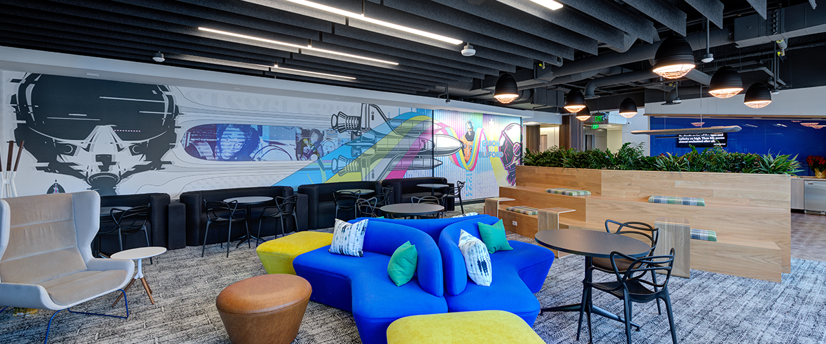 A bright and open collaboration space with featured artwork on one wall, blue and yellow bright colored couches, and black chairs for seating.