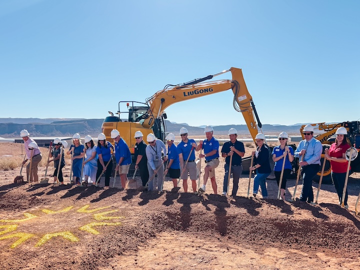 City, county, and state officials stand in a row with shovels at the groundbreaking ceremony for Utah’s first air traffic control tower outside the Wasatch Front at St. George Regional Airport. A yellow excavator is in the background.