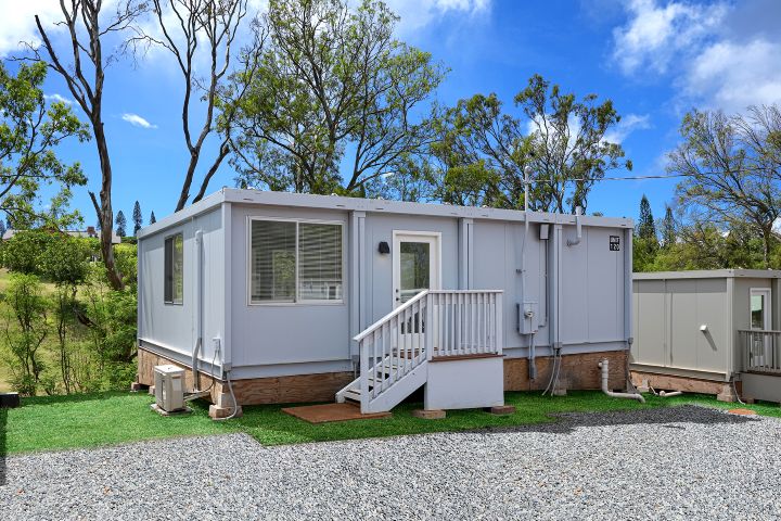 Modular emergency housing unit with white exterior, front porch, and gravel driveway, set in a green, tree-lined area in Lahaina, Hawaii. p-hero-w-heading__img
