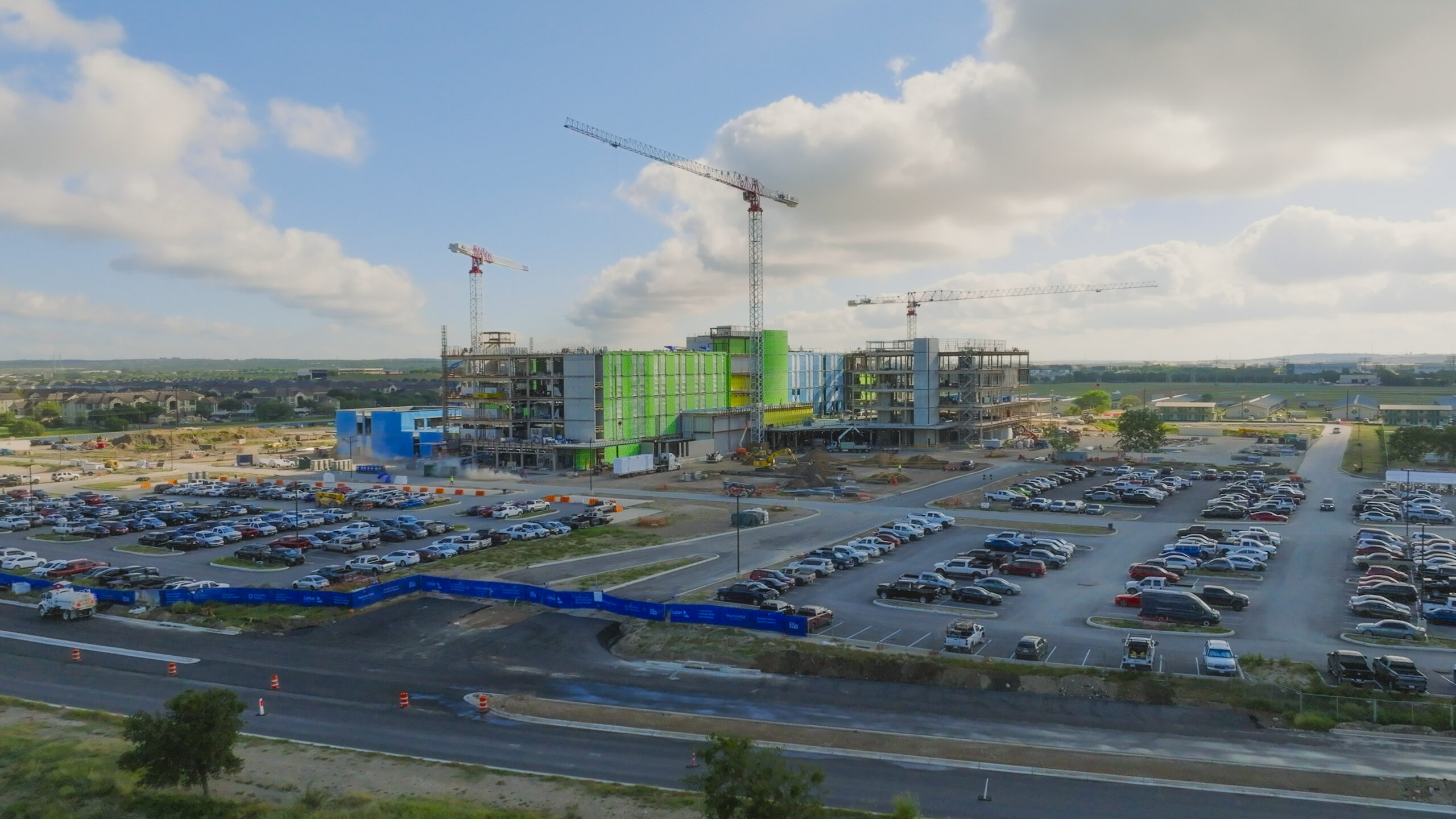 Aerial view of the University Health Retama Community Hospital under construction. The multi-story building features cranes, partially completed exterior walls with green and blue sheathing, and active construction equipment around the site. Surrounding the hospital are dirt lots, vehicles, and nearby structures.