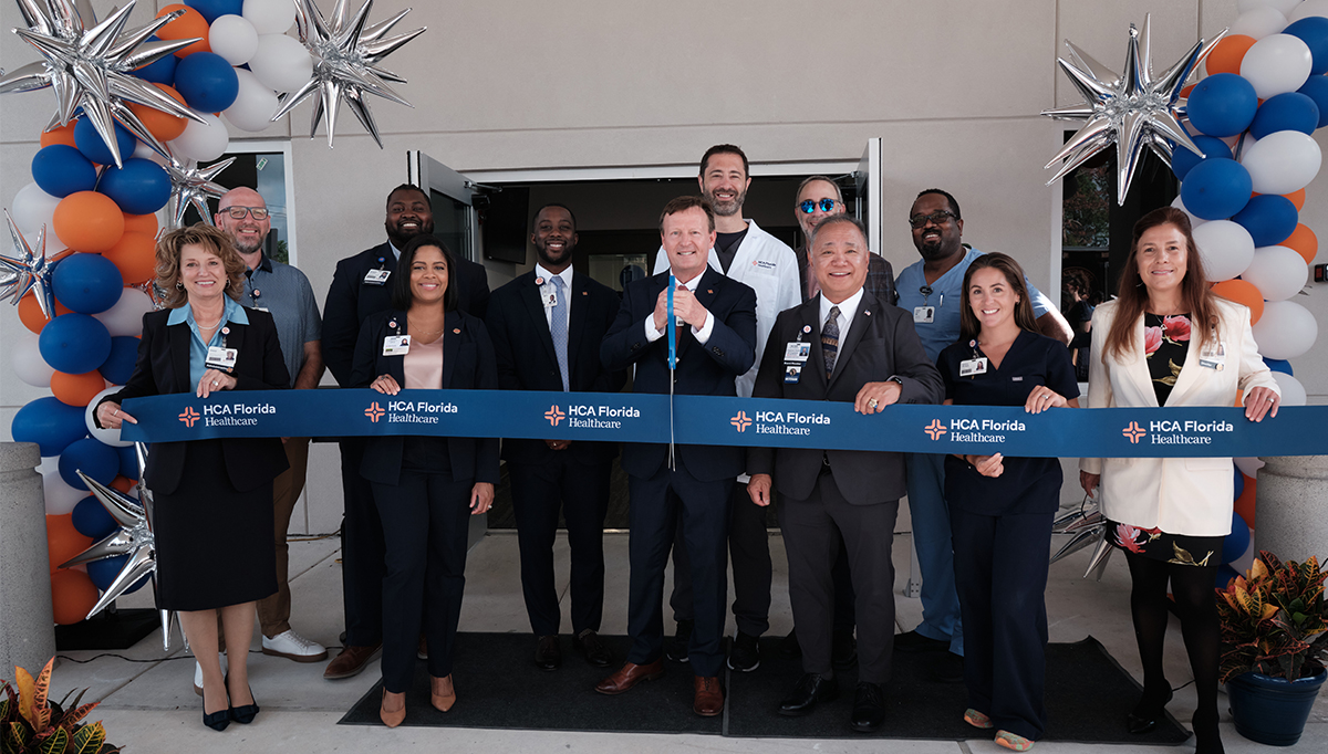 Crowd of men and women cut ribbon in front of double doors surrounded by white, blue, and orange balloons.